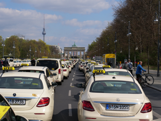 Lange Reihe von Taxis entlang einer belebten Straße in Berlin, Deutschland, mit Fahrradfahrern und Füttern auf dem Gehweg, flankiert von Laternen und Bäumen und Gebäuden, einem Bogen und einem Turm im Hintergrund bei einem bewölktem Himmel.