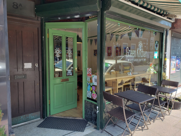 Außensitzbereich des Green Kitchen Vegan Cafe mit Tischen, Stühlen, Pflanzen, einer Matte und einer Glastür mit Postern, der zu einem Innenraum mit Tischen, Stühlen und Deckenleuchten führt.