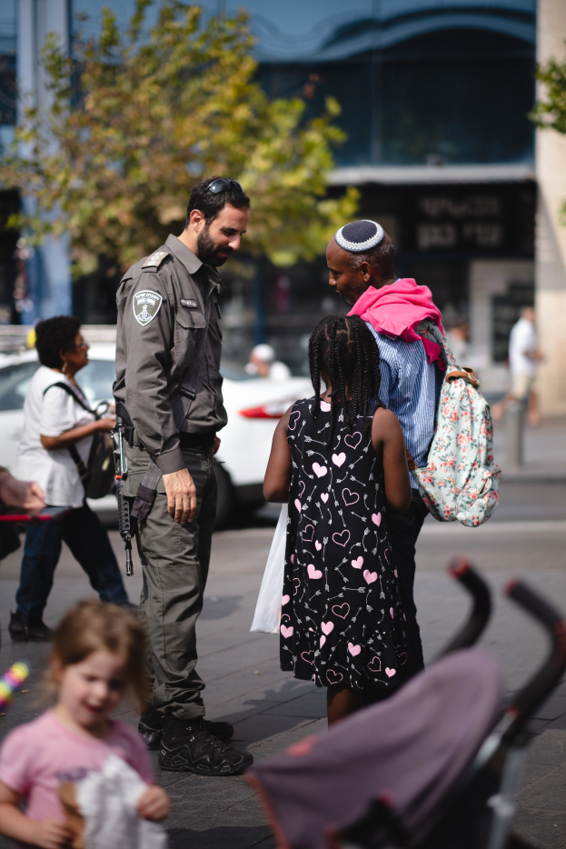 Ein Polizeibeamter spricht mit einer Gruppe von Menschen auf der Straße, mit einem Kinderwagen im Vordergrund und Fahrzeugen, Bäumen und einem Gebäude im Hintergrund.