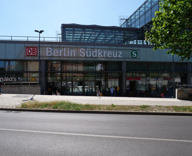 Großes gläsernes Berliner Südkreuz-Bahnhof mit Text an seiner Fassade, umgeben von Straßenlaternen, Lampen, Fahrzeugen, Fußgängern, Fahrrädern, Bäumen und einem klaren blauen Himmel.