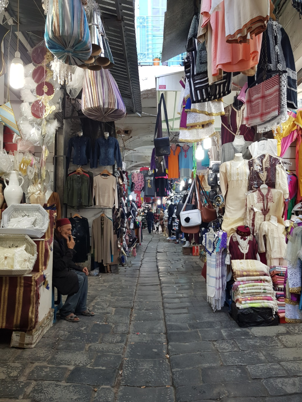 Ein belebter Straßenmarkt in der Altstadt von Jerusalem mit Ständen, die Kleidung, Taschen, Lampen und andere Gegenstände verkaufen, umgeben von Menschen, die durch die Stände schlendern und eine Gebäudefront im Hintergrund.
