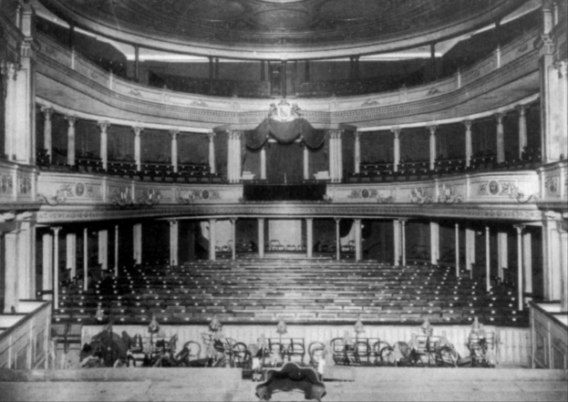 Altes Schwarz-Weiß-Foto des Theatre Royal Auditoriums in London, das Sitzreihen, Treppen, Geländer, Säulen, Lampen und einen Kronleuchter mit einigen sitzenden und stehenden Menschen zeigt.