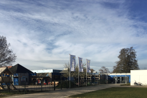 Großes Gebäude mit Metallzaun, Rasenfläche, Weg, Bäumen, Pfählen, Bannern, einem geparkten Fahrrad, nahen Häusern und einem bewölkten Himmel, identifiziert als der neue Spielplatz der Schule.