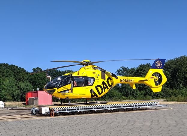 Ein gelber Hubschrauber steht auf einem Flughafenrollfeld umgeben von Bäumen und einem klaren blauen Himmel, mit verschiedenen Gegenständen, die mit dem Hubschrauber in Zusammenhang stehen, herumstreut.