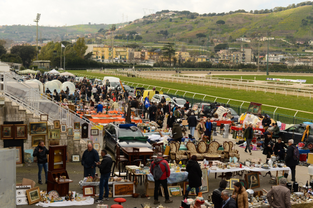 Eine große Gruppe von Menschen auf einem Outdoor-Flohmarkt mit Tischen, auf denen Gegenstände wie Foto Rahmen und Stühle ausgelegt sind, geparkte Fahrzeuge, Geländer, Stufen, Bäume, Gebäude, Laternenpfähle, Hügel und ein bewölkter Himmel.