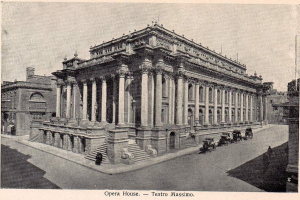 Ein altes Schwarzweißfoto des Teatro Massimo Opernhauses in Rom, Italien, das ein großes Gebäude mit Säulen, Stufen und Bögen zeigt, sowie einige Menschen und Karren auf der Straße davor und Text am unteren Rand des Bildes.