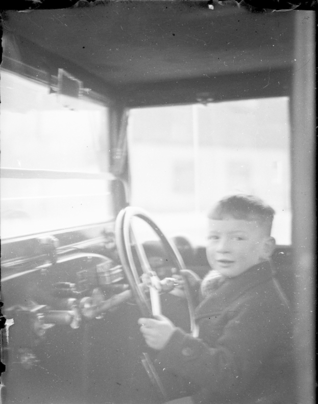 Ein junger Junge in Schwarz-Weiß sitzt auf dem Fahrersitz eines Autos und hält das Lenkrad fest mit beiden Händen.