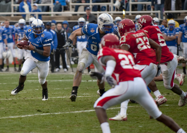 Fußballspieler in weißem Trikot mit dem Ball läuft, verfolgt von Gegenspielern in dunklen Trikots und Helmen, auf einem Rasenfeld mit Zuschauern und Geländern im Hintergrund.