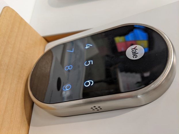 Nahaufnahme einer Yale Smart Lock-Vorrichtung auf einem weißen Tisch mit einem hölzernen Objekt links daneben.