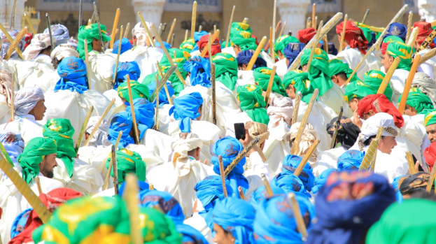 Eine große Gruppe von Menschen in farbenfrohen Kostümen marschiert eine Straße entlang, jeder hält einen Stock in der Hand, mit einem Gebäude im Hintergrund.