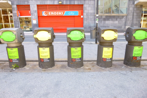 Reihe von Parkautomaten entlang einer Straße mit einem Gebäude mit Glasfenstern und einer Rolllade mit Text sowie einem nahen Müllcontainer.