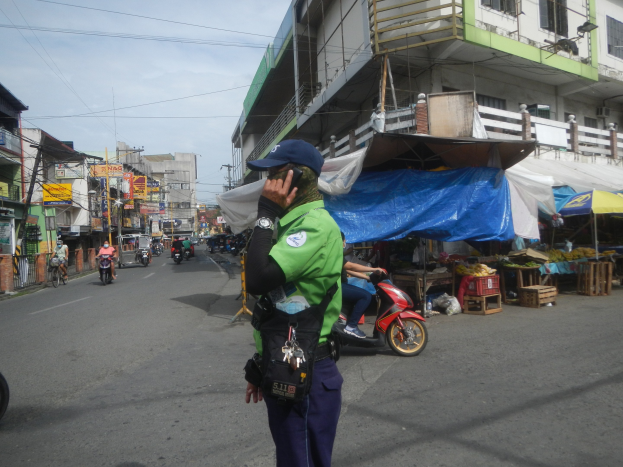 Polizist in grüner und blauer Uniform mit blauer Mütze auf einer Straße stehend, der auf einem Handy spricht, mit Fahrzeugen und Marktständen im Hintergrund und einem Gebäude mit Fenstern im bewölkten Himmel.