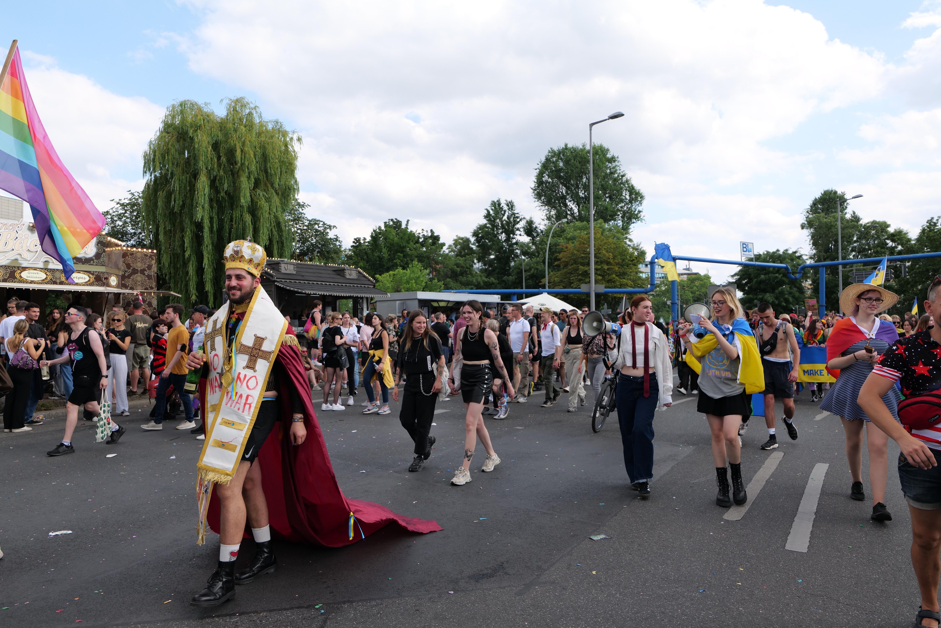 Eine Gruppe von Menschen, die bei der Gay Pride Parade 2018 marschieren und eine Regenbogenflagge sowie Musikinstrumente tragen, während einige Mützen tragen, vor dem Hintergrund von Laternenpfählen, Bäumen, Schuppen und einem bewölkten Himmel.