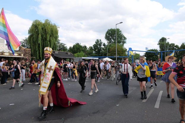 Eine Gruppe von Menschen, die bei der Gay Pride Parade 2018 marschieren und eine Regenbogenflagge sowie Musikinstrumente tragen, während einige Mützen tragen, vor dem Hintergrund von Laternenpfählen, Bäumen, Schuppen und einem bewölkten Himmel.