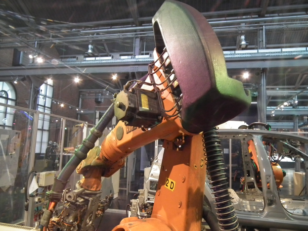Ein Roboterarm arbeitet an einem Auto in einer industriellen Umgebung mit Metallstangen, Beleuchtung und Hintergrundmaschinen, getrennt durch eine Glaswand.