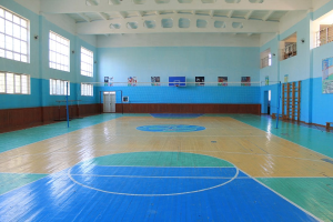Große Turnhalle mit blau-weißem Boden, Basketballfeld, Netz, Stangen, Tür, Fenstern, Wandplakaten, Geländer und Dach.