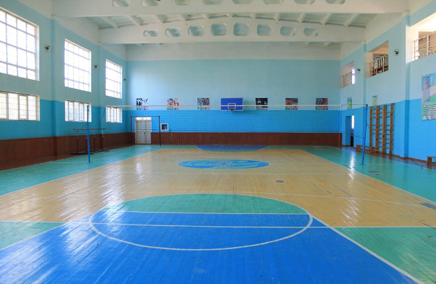 Große Turnhalle mit blau-weißem Boden, Basketballfeld, Netz, Stangen, Tür, Fenstern, Wandplakaten, Geländer und Dach.