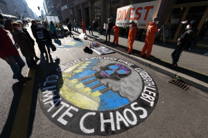 Eine Gruppe von Menschen steht auf einer Straße, einige tragen Masken und halten Schilder mit der Aufschrift "Klimachaos", die gegen die Regierungsschließung protestieren, mit verstreuten Flaschen und Gebäuden im Hintergrund unter einem klaren blauen Himmel.