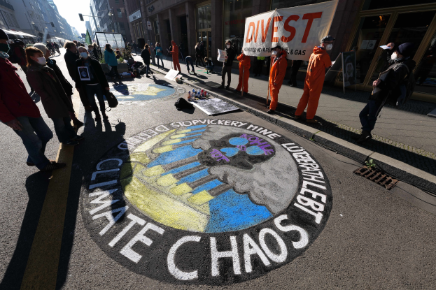 Eine Gruppe von Menschen steht auf einer Straße, einige tragen Masken und halten Schilder mit der Aufschrift "Klimachaos", die gegen die Regierungsschließung protestieren, mit verstreuten Flaschen und Gebäuden im Hintergrund unter einem klaren blauen Himmel.