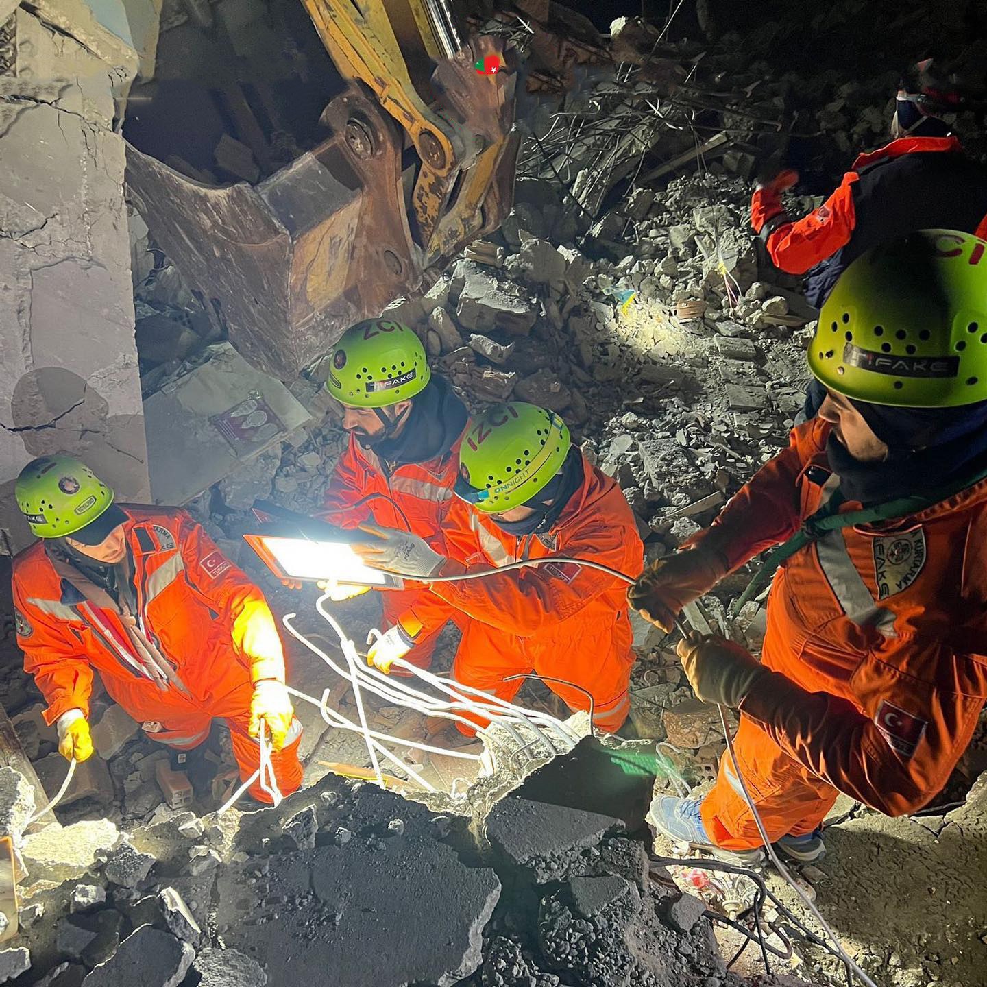 Eine Gruppe von Menschen in orangefarbenen Uniformen und grünen Helmen bei der Arbeit an Schutt von einem eingestürzten Gebäude, mit einem Kran im Hintergrund, höldend Rettungsgeräte und -materialien.
