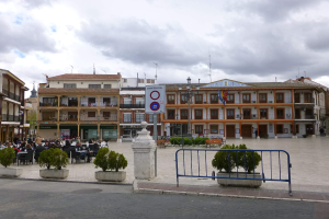 Ein belebter Stadtplatz mit sitzenden und stehenden Menschen, Topfpflanzen, Metallabsperrungen, einem Schild an einem Pfahl, Straßenlaternen mit Flaggen, Gebäuden mit Fenstern und einem bewölkten Himmel.