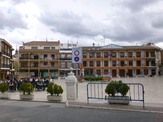 Ein belebter Stadtplatz mit sitzenden und stehenden Menschen, Topfpflanzen, Metallabsperrungen, einem Schild an einem Pfahl, Straßenlaternen mit Flaggen, Gebäuden mit Fenstern und einem bewölkten Himmel.