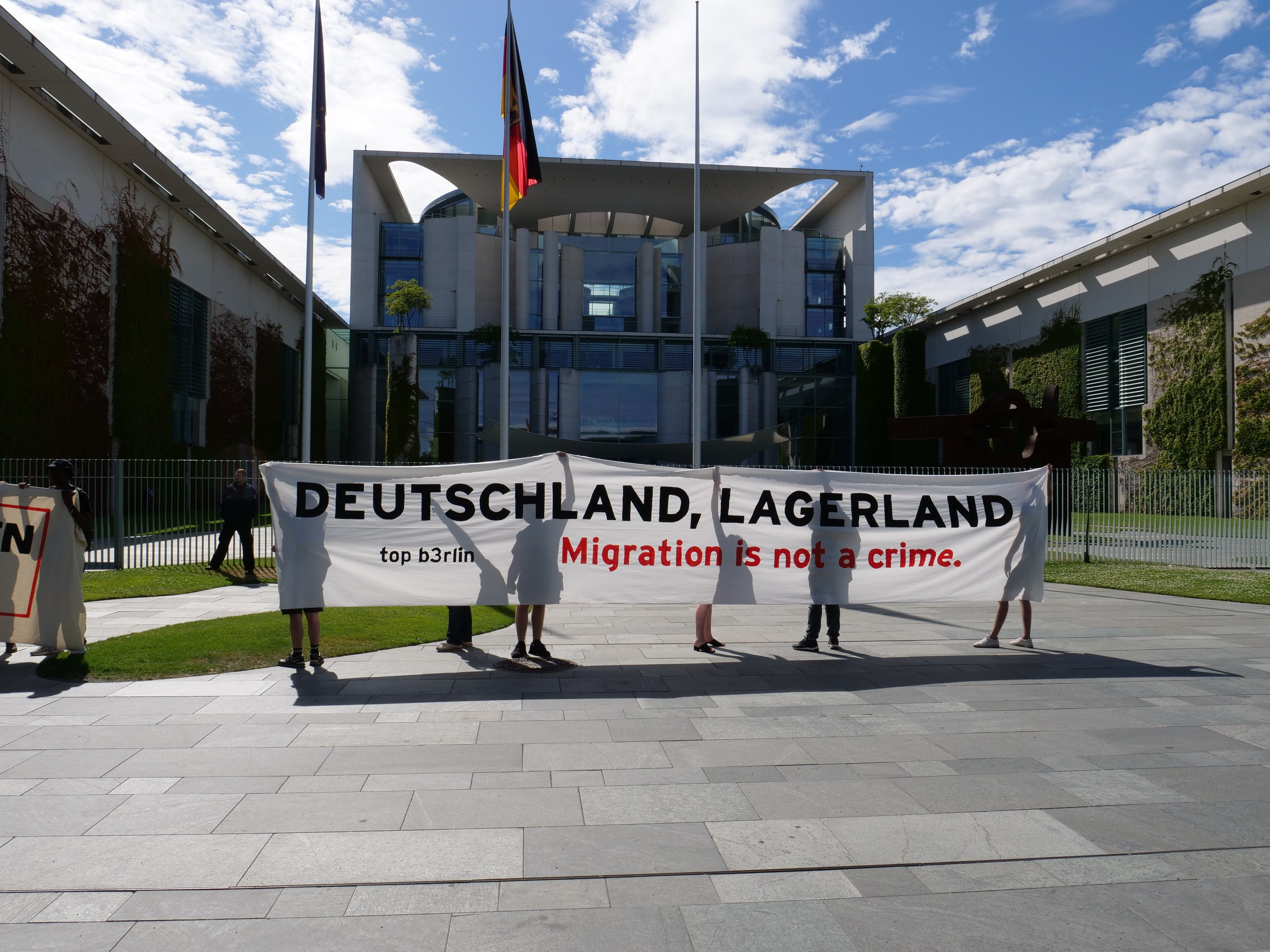 Eine Gruppe von Menschen mit einer Fahne, auf der "Deutschland, Lagerland Migration ist kein Verbrechen" steht, vor einem Gebäude mit Fenstern, umgeben von Gras, einem Metallzaun, Pflanzen, Bäumen und Fahnenmasten vor einem bewölkten Himmel.