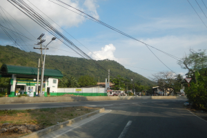 Tankstelle an einer Straße mit Strommasten, Kabeln, Schildern, Bäumen, Gebäuden, Fahrzeugen, Hügeln und bewölktem Himmel im Hintergrund.