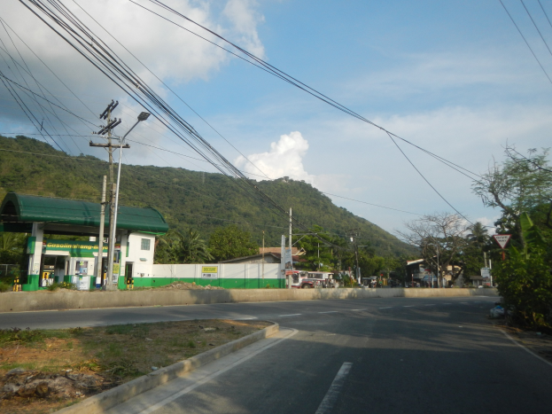 Tankstelle an einer Straße mit Strommasten, Kabeln, Schildern, Bäumen, Gebäuden, Fahrzeugen, Hügeln und bewölktem Himmel im Hintergrund.