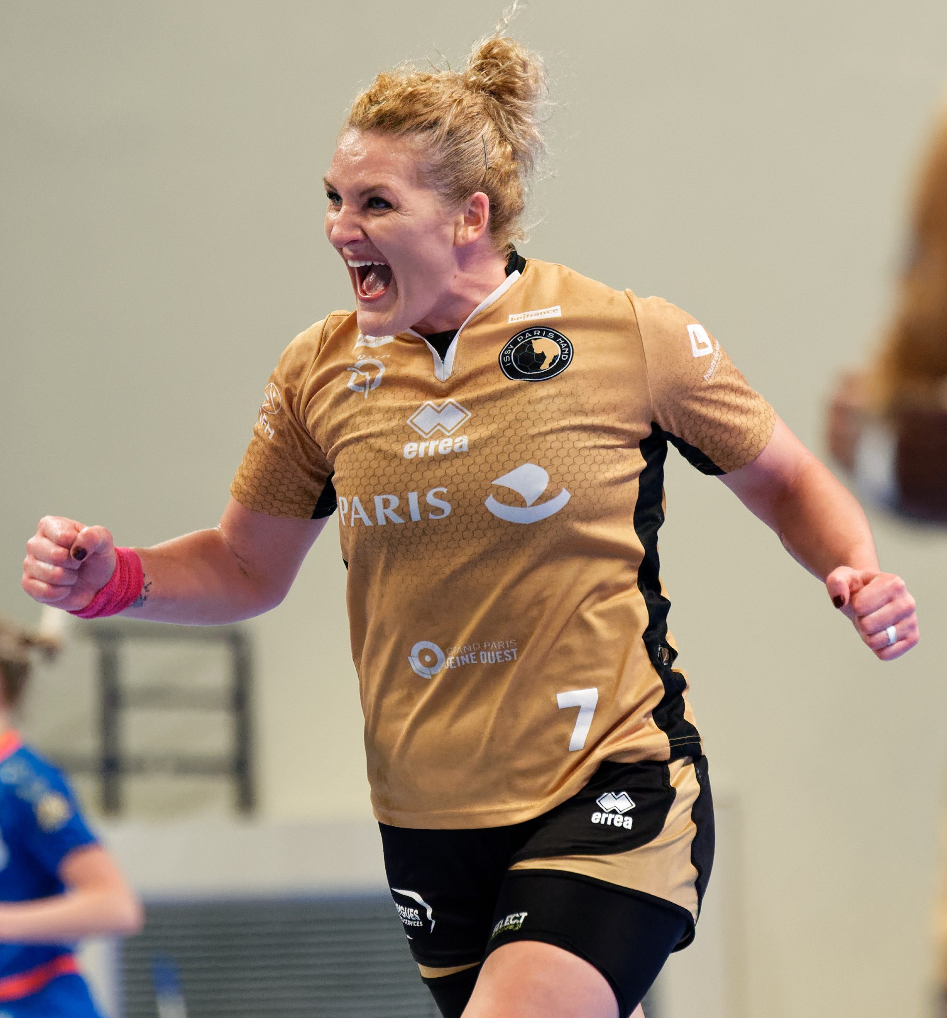 Eine Frau in braunem T-Shirt und schwarzen Shorts rennt und feiert auf einem Futsalplatz, mit zwei anderen Frauen im Hintergrund.