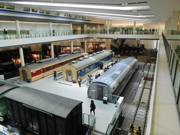 Ein großer Museumsraum mit verschiedenen Zügen in unterschiedlichen Größen und Farben, bei dem Menschen die Ausstellungsstücke beobachten und Deckenleuchten den Raum erhellen.