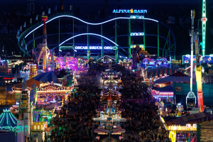 Eine große Menschenmenge bei einem nächtlichen Karneval, beleuchtet von bunten Lichtern, umgeben von Vergnügungsgeräten, Pfählen, Fahnen und Namensschildern, mit einem dunklen Hintergrund, der eine festliche Atmosphäre schafft.
