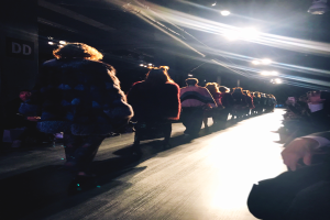 Eine Gruppe von Menschen läuft auf einem Laufsteg bei einer Modenschau, beleuchtet von hellen Scheinwerfern, mit Zuschauern auf beiden Seiten.