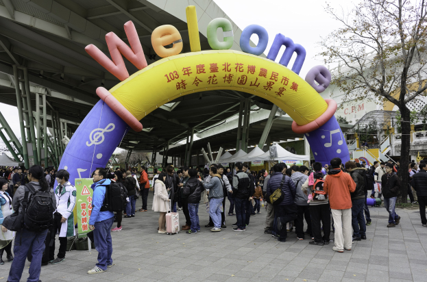 Eine Gruppe von Menschen vor einem Willkommensbogen bei einem Outdoor-Event mit Zelten, Bäumen und Gebäuden unter einem klaren blauen Himmel, einige tragen Taschen.