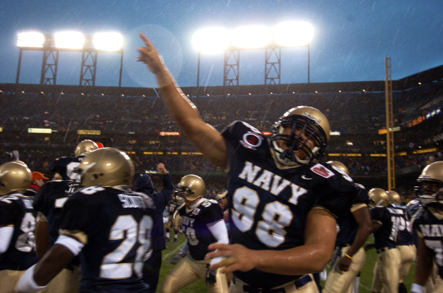 Navy-Football-Spieler feiern Touchdown im Regen, tragen Helme und Uniformen auf einem Rasenfeld mit Zuschauern und Stadionbeleuchtung im Hintergrund.
