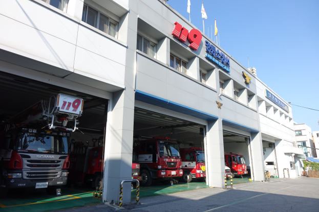 Feuerwache mit mehreren Feuerwehrautos vor der Tür, mit Fenstern, Säulen, Namensschildern und Fahnen im Vordergrund vor anderen Gebäuden und einem klaren blauen Himmel.