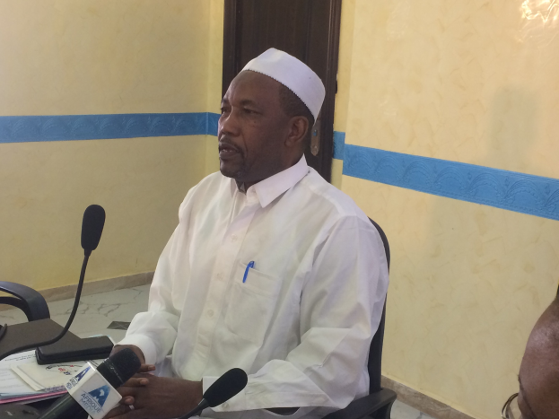 Ein Mann in einem weißen Kleid und Hut sitzt an einem Tisch mit Mikrofonen, spricht, während Papiere und Gegenstände auf dem Tisch sichtbar sind, mit einem anderen Mann mit Brille zu seiner Rechten, in einem Raum mit einer Wand und einer Tür im Hintergrund.