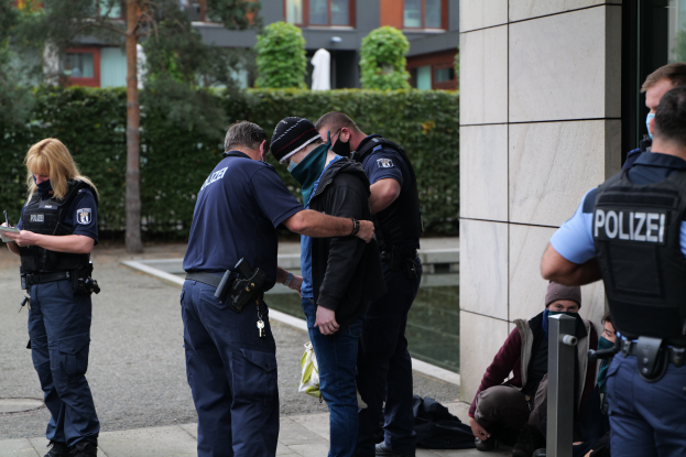 Eine Gruppe von Polizeibeamten steht zusammen vor einem Gebäude, mit einer Frau, die ein Buch und einen Stift hält, während einige Menschen stehen und andere auf dem Boden sitzen, mit Pflanzen, Bäumen und Gebäuden im Hintergrund.