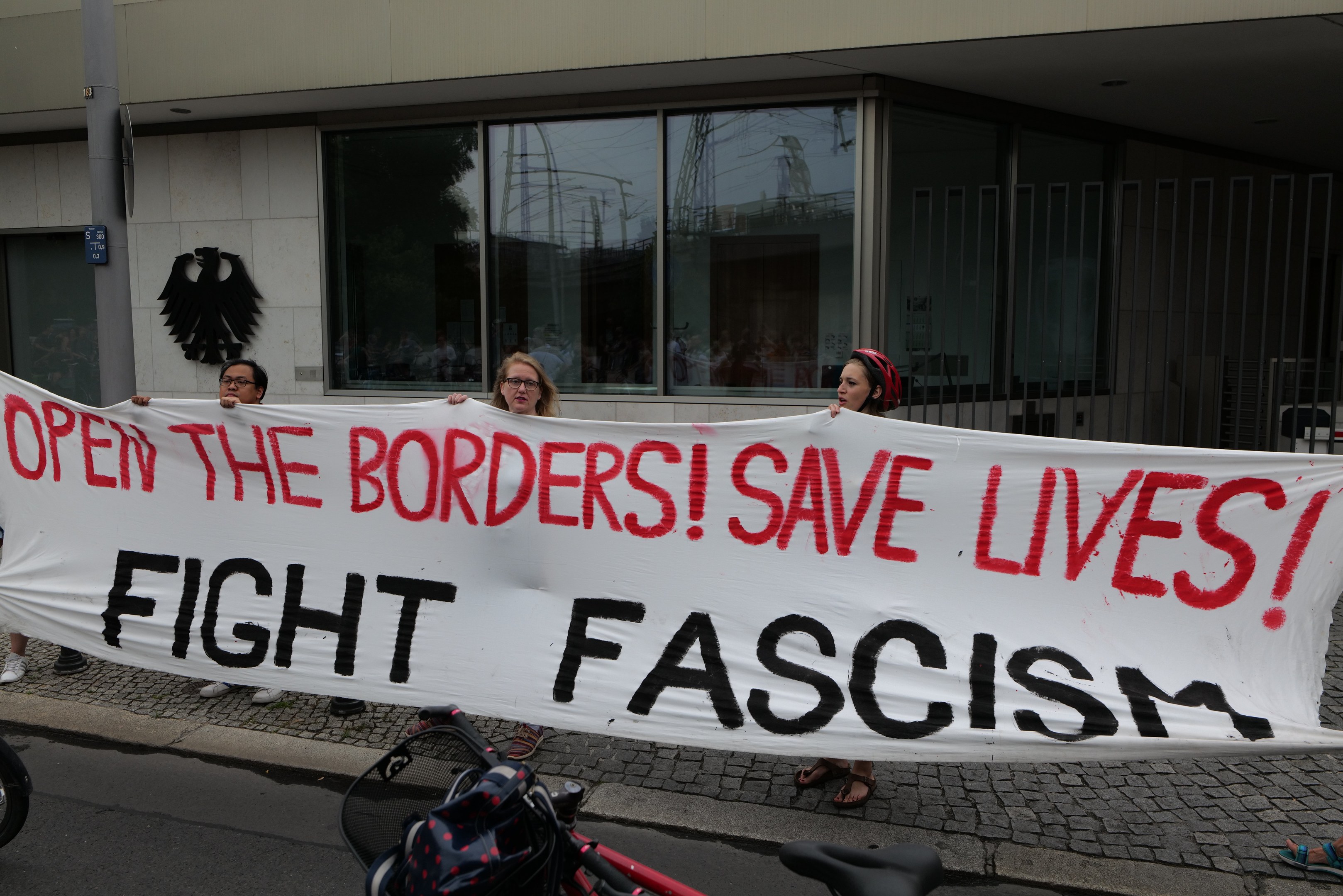 Menschen mit einem Transparent mit der Aufschrift "Öffnet die Grenzen, rettet Leben, kämpft gegen Faschismus" vor einem Gebäude mit Glasfenstern und einem Pfahl, mit einem Fahrrad und einer Tasche auf der Straße.