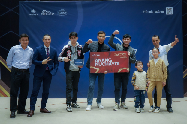 Gruppe von Menschen auf einer Bühne mit einer Tafel, die "Akang Kuchaydi - Zakovat Quiz" zeigt, im Hintergrund ein Banner und ein Barcode.