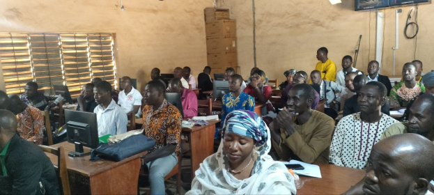 Eine Gruppe von Menschen sitzt an Tischen in einem classroom mit Monitoren, Taschen, Büchern und Papieren auf den Tischen, ein Fenster auf der linken Seite und Kartons und einen Fernseher an der Wand im Hintergrund, nimmt an einem Techniktraining teil.