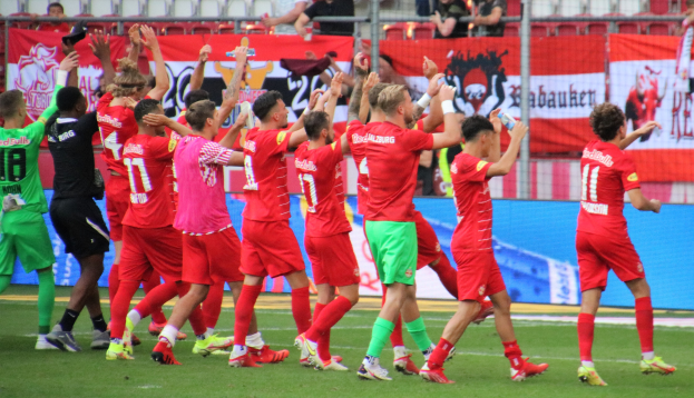Gruppe von Fussballspielern, die ein Tor feiern, auf einem Feld mit Flaschen oder klatschenden Händen, mit einem Zaun, Bannern, Metallstangen, Stühlen und einem Dach mit Deckenleuchten im Hintergrund.