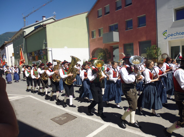 Eine Gruppe von Menschen in traditioneller bayrischer Tracht, die auf der Straße Musikinstrumente spielen und dabei durch eine Straße mit Gebäuden gehen, einige halten Fahnen, im Hintergrund ein Hügel und ein klarer blauer Himmel.