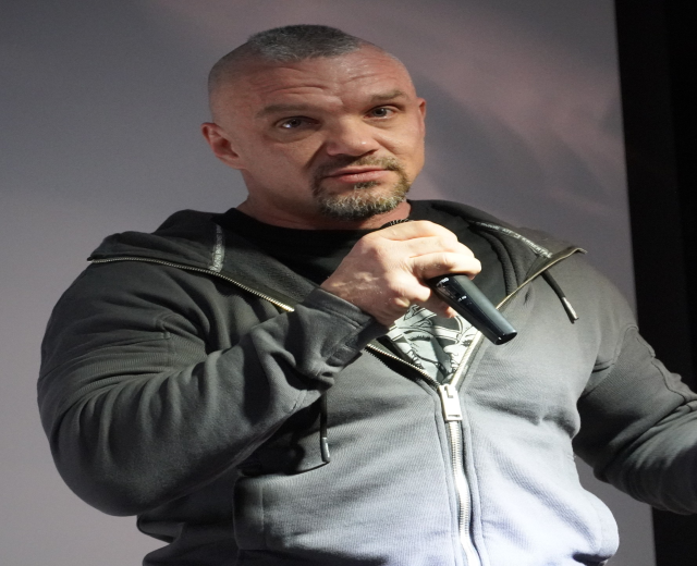 Ein Mann in einer schwarzen Kapuzenjacke und einem schwarzen Anzug mit einem wei├čen Hemd, der ein Mikrofon h├Ąlt und selbstbewusst mit zurückgek├Ąmmtem Haar vor einer Wand steht.