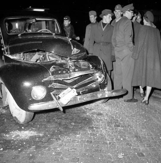 Schwarz-weißes Bild eines schwer beschädigten Autos am Straßenrand mit eingedellter Motorhaube und zersplitterten Fenstern, umgeben von einer Gruppe von Menschen, die die Szene begutachten.