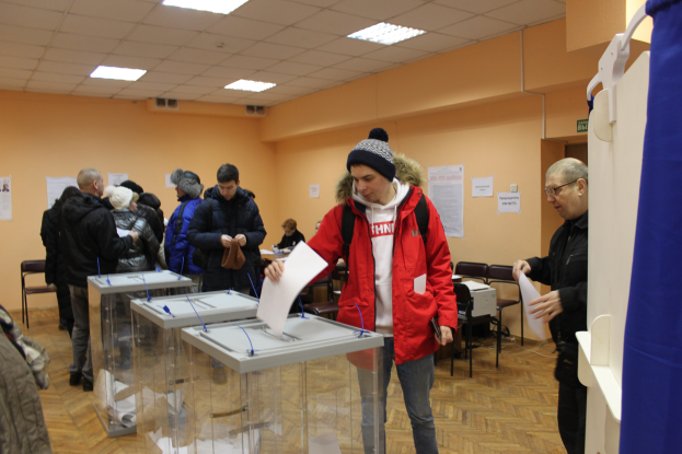 Eine Gruppe von Menschen steht um eine Wahlkabine in einer Wahllokal, einige halten Papiere, mit einem blauen Vorhang und weißem Objekt auf der rechten Seite, Stühle, Tische, Papiere an der Wand und Deckenleuchten im Hintergrund.
