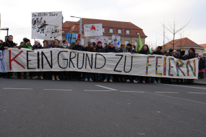 Gruppe von Menschen mit einem Transparent mit der Aufschrift 'Kein Grund zu Feiern' bei einer Demonstration gegen die Sparpolitik der deutschen Regierung, im Hintergrund Straßeninfrastruktur und Gebäude unter einem klaren Himmel.
