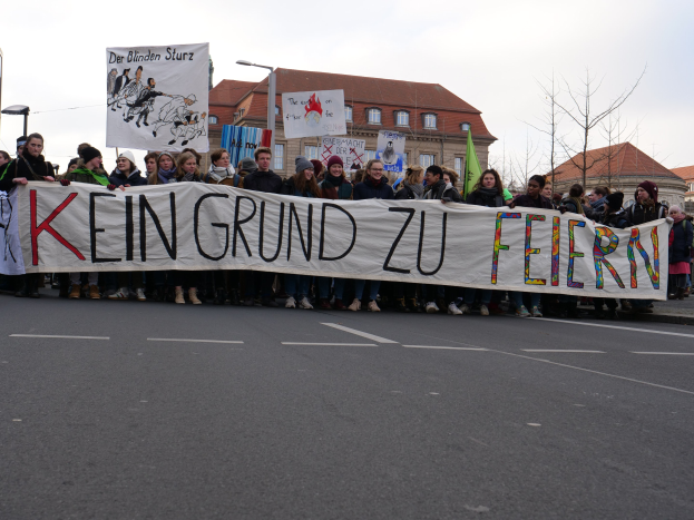 Gruppe von Menschen mit einem Transparent mit der Aufschrift 'Kein Grund zu Feiern' bei einer Demonstration gegen die Sparpolitik der deutschen Regierung, im Hintergrund Straßeninfrastruktur und Gebäude unter einem klaren Himmel.