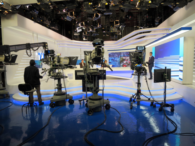 Ein TV-Studio mit mehreren Kameras auf dem Boden, einer sitzenden Person im Vordergrund und einigen stehenden Personen im Hintergrund, mit an der Decke montierten Scheinwerfern für eine Live-Broadcast-Aufstellung.