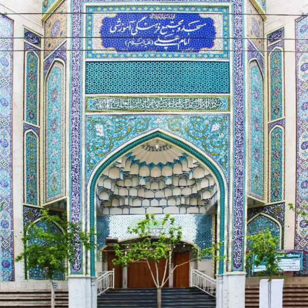 Blau-weiß gekachelte Wand mit detaillierten Designs und Text am Eingang eines Gebäudes, mit Stufen, Geländern und Bäumen im Vordergrund, sowie einem Informationsschild über Isfahan, Iran.